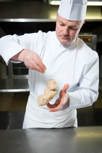 Un chef cuisinier en toque et veste blanche assaisonne un bloc de foie gras qu'il tient dans ses mains.