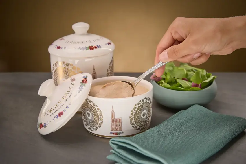 Une main prélève une cuillère de pâté dans une terrine décorée, à côté d'une salade verte.